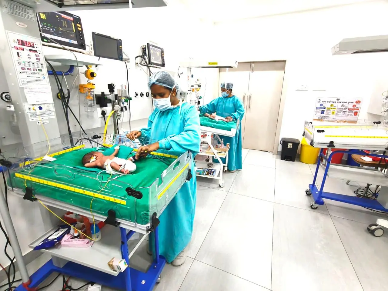 Newborn in a neonatal incubator being cared for by a nurse in blue scrubs in a NICU, with monitors and equipment nearby into the scene.
