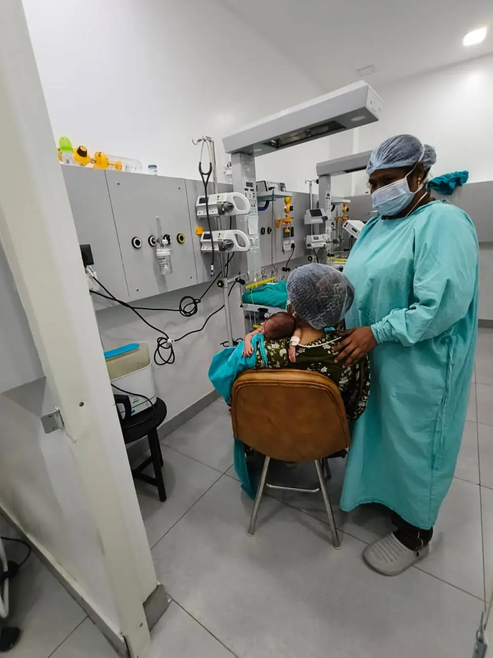 Nurse in teal scrubs and mask standing beside a seated mother holding a newborn, in a hospital room with medical panels and equipment in the background.