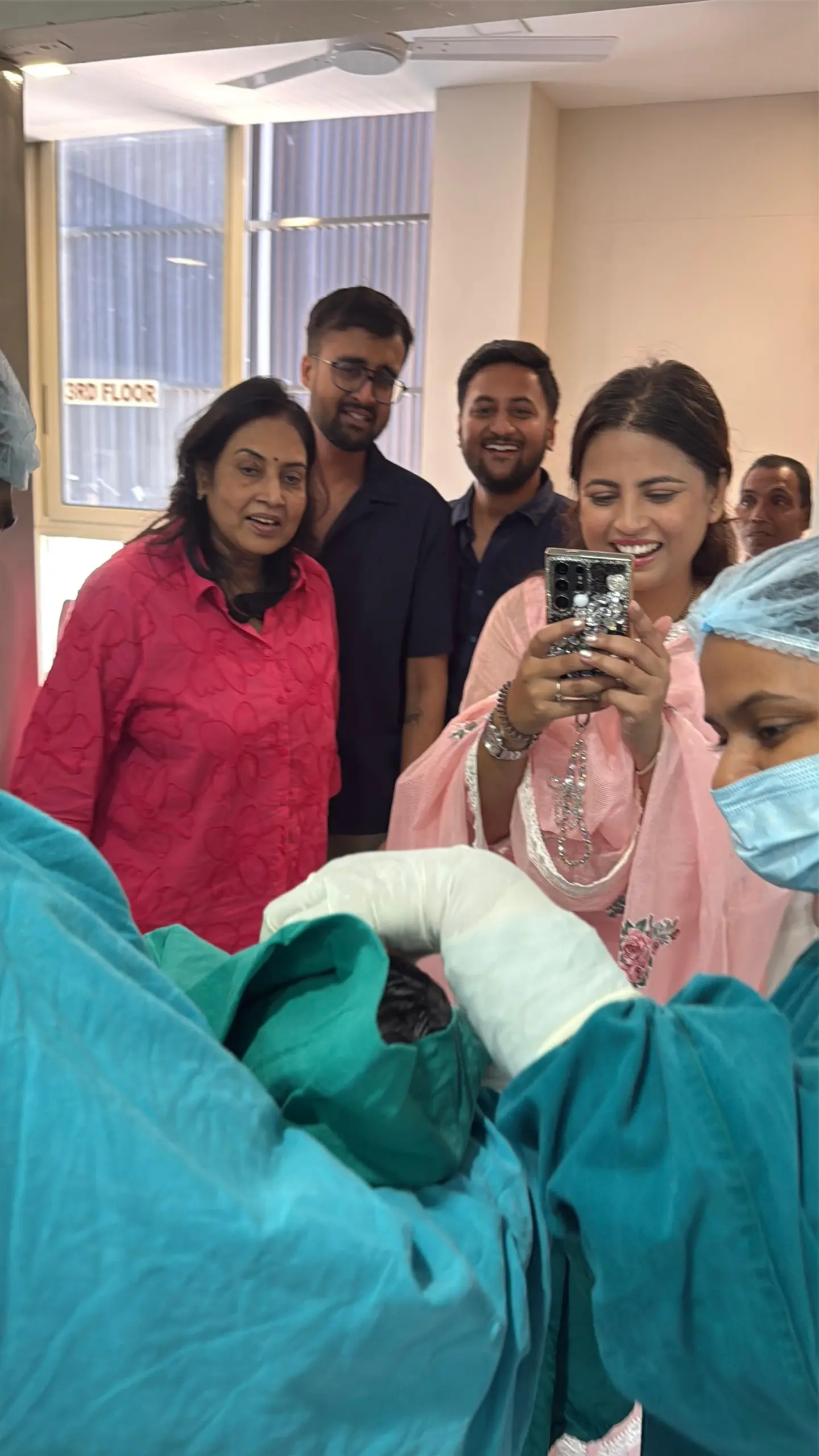 Nurse in teal scrubs and mask standing beside a seated mother holding a newborn, in a hospital room with medical panels and equipment in the background.