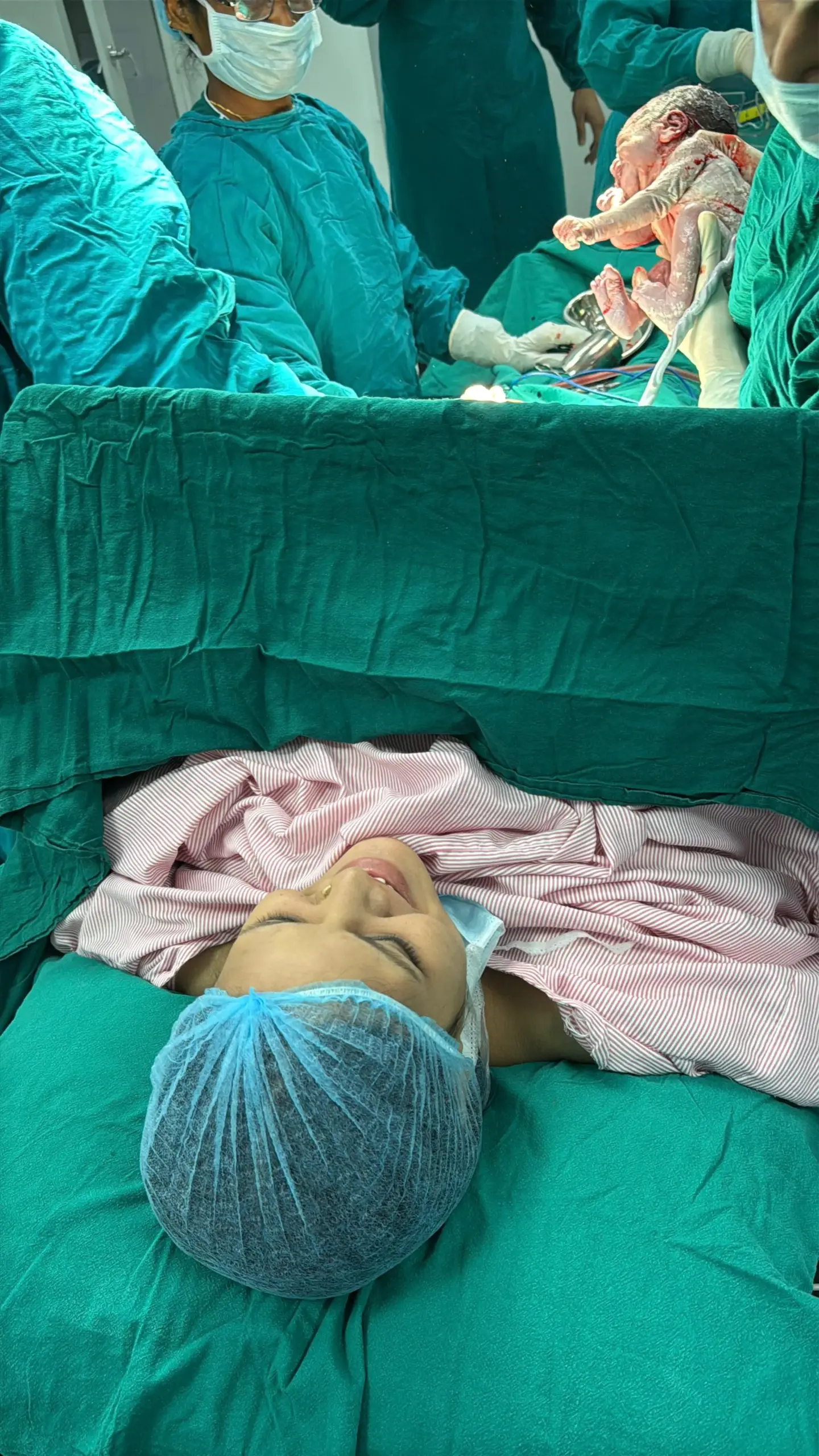 Nurse in teal scrubs and mask standing beside a seated mother holding a newborn, in a hospital room with medical panels and equipment in the background.